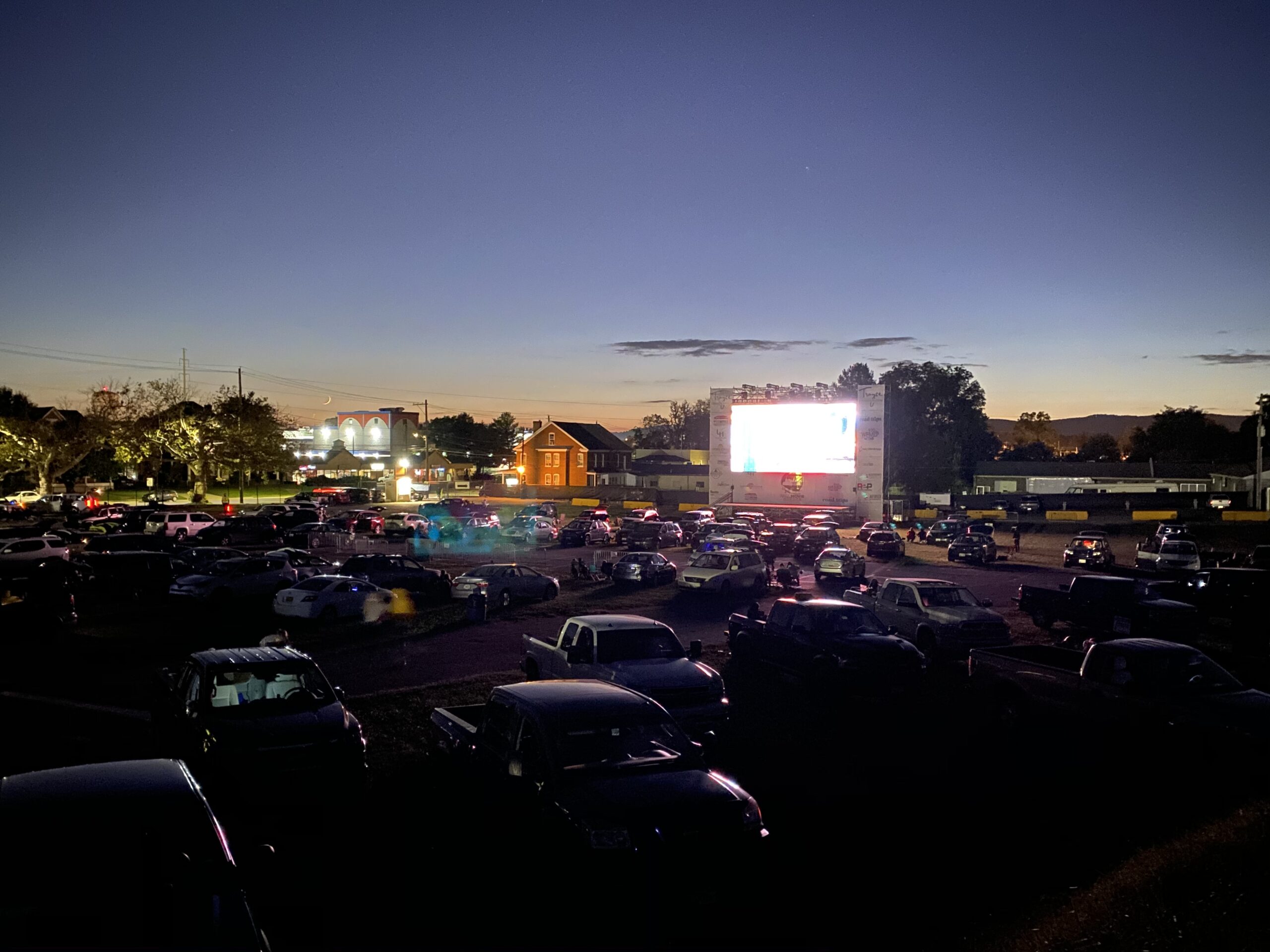 Dusk scene of a large outdoor movie screen glowing white, surrounded by a packed parking lot of cars and trucks under a twilight sky