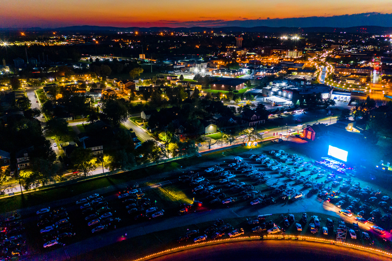 Aerial night view of a town with illuminated streets, rows of parked cars, and a large glowing screen near a sunset horizon